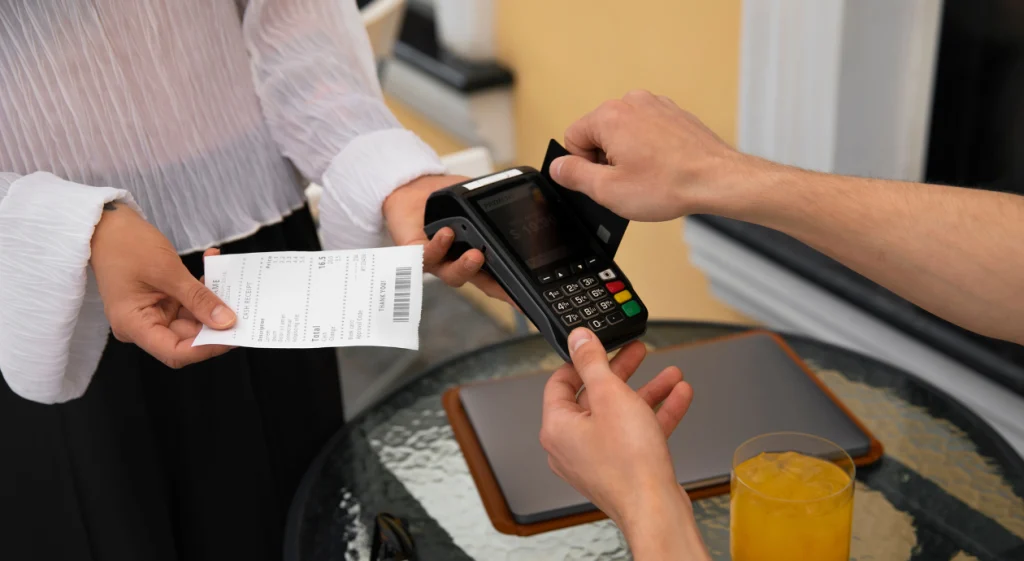 Close-up de uma transação de pagamento: mão inserindo cartão na maquininha e outra segurando um cupom fiscal