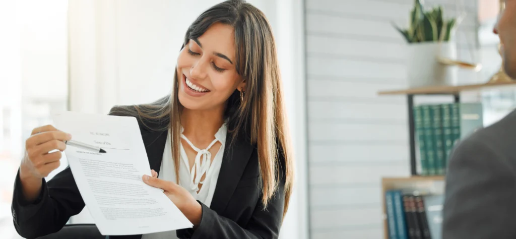 Mulher de negócios sorridente em blazer, apontando para um documento com caneta, conversando com um colega.
