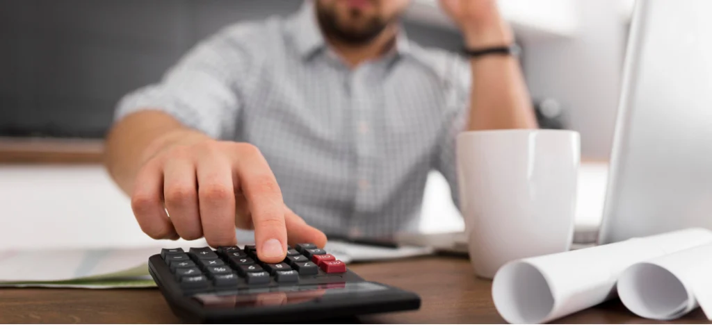 Mão em primeiro plano usando uma calculadora sobre uma mesa com papéis, xícara e notebook, sugerindo cálculo de custos.