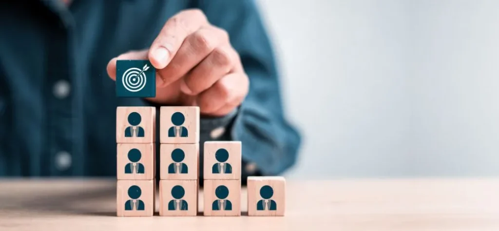 Mão colocando um bloco de alvo no topo de uma pirâmide de blocos com ícones de pessoas. Foco no cliente/Liderança.