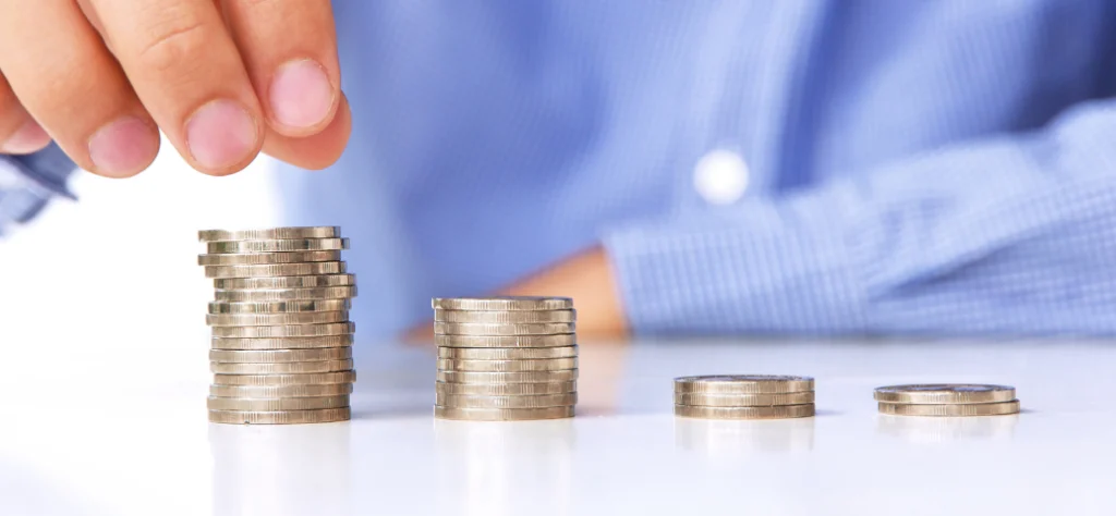 Mão colocando uma moeda no topo de uma pilha de moedas, com outras pilhas menores em uma superfície branca.