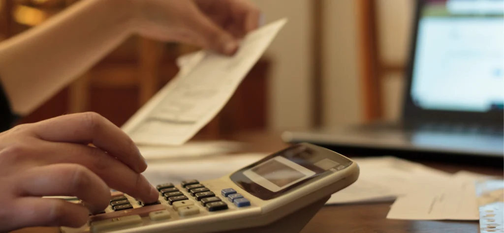 Pessoa digitando em uma calculadora, revisando documentos e usando um laptop ao fundo. Gestão de finanças/Fluxo de caixa.