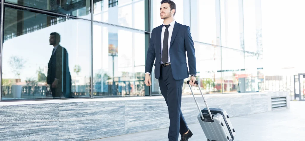 Homem de terno, com mala de rodinhas, caminhando para fora de um edifício de vidro em um dia ensolarado.