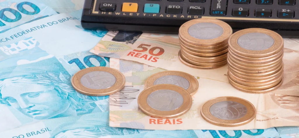 Pilhas de moedas de Real e notas de R$ 50 e R$ 100 ao lado de uma calculadora, representando impostos e economia
