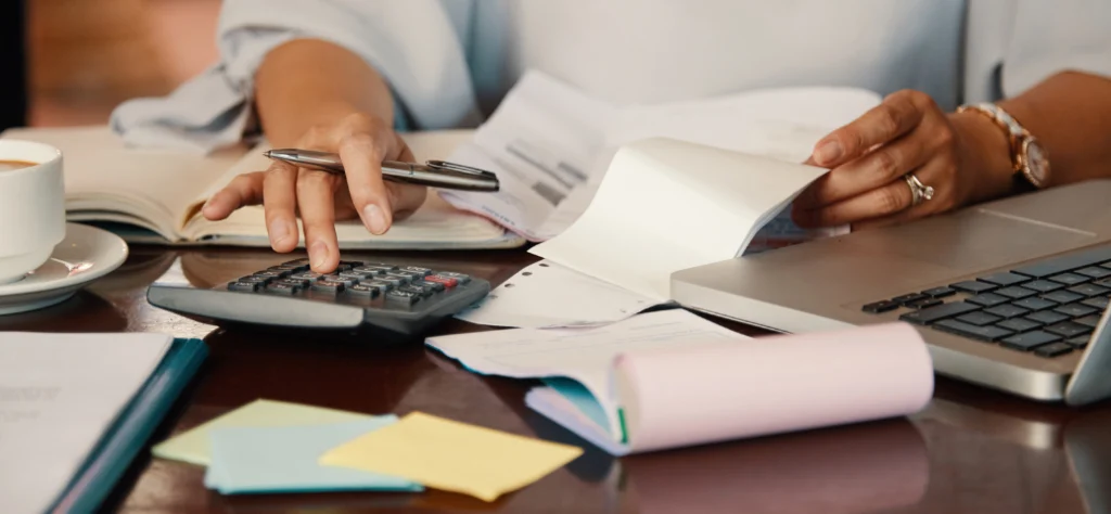 Pessoa com caneta e calculadora em uma mesa com documentos e laptop, fazendo cálculos de folha ou impostos.