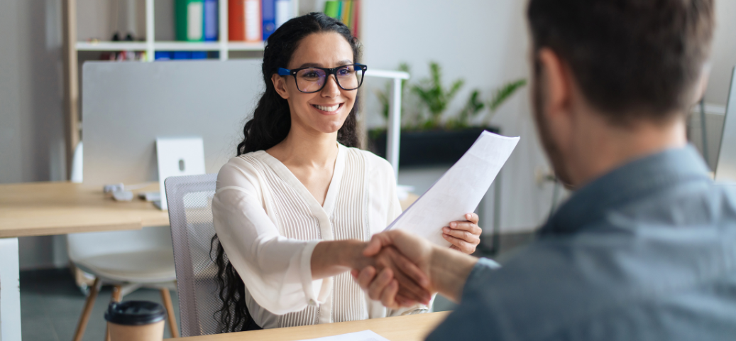 Mulher sorridente de óculos apertando a mão de um homem em um escritório, segurando um documento. Contratação/Entrevista.