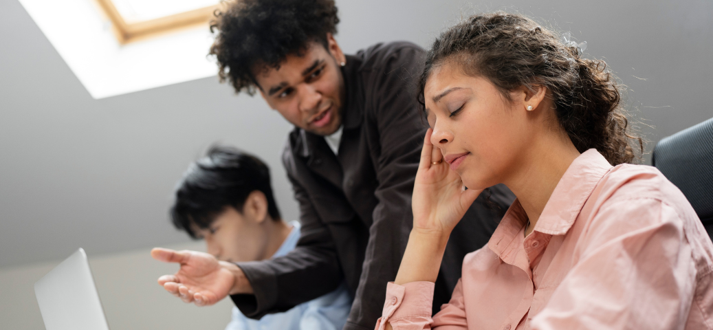 Homem em camisa escura gesticula, apontando para um notebook enquanto uma mulher em camisa rosa segura a cabeça parecendo estressada.