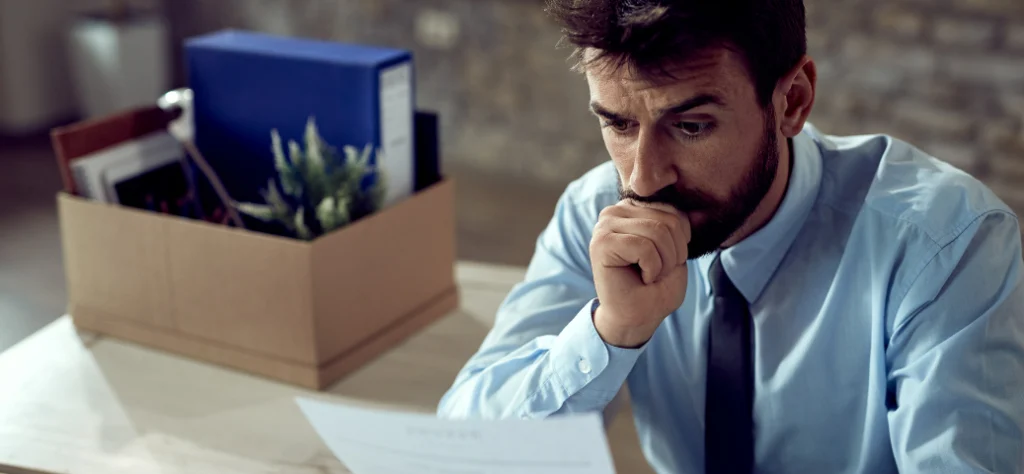 Homem de terno e gravata, com expressão preocupada, olhando para um documento ao lado de uma caixa de demissão. Multas/Problemas.