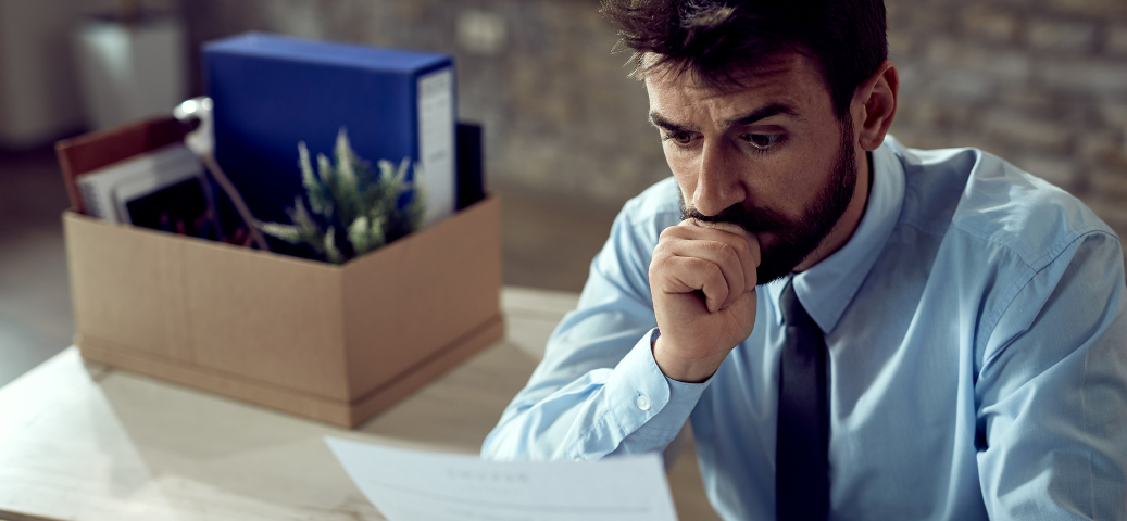 Homem de terno e gravata, com expressão preocupada, olhando para um documento ao lado de uma caixa de demissão. Multas/Problemas.