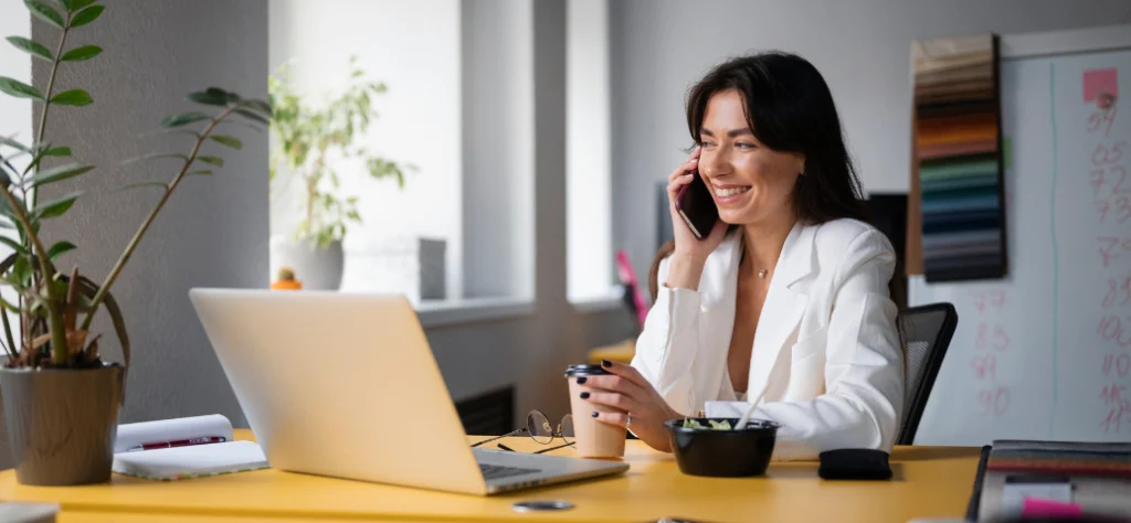 Mulher sorridente, com blazer branco, ao telefone em frente a um notebook em uma mesa amarela com um copo de café e uma tigela de comida.