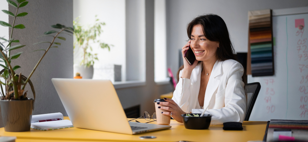 Mulher sorridente, com blazer branco, ao telefone em frente a um notebook em uma mesa amarela com um copo de café e uma tigela de comida.