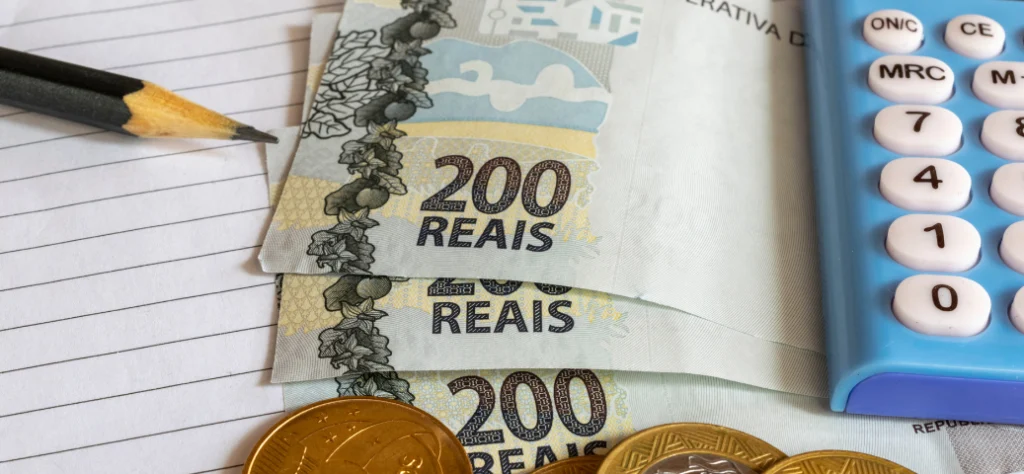 Três notas de R$ 200, moedas, um lápis preto e uma calculadora azul sobre um papel pautado.