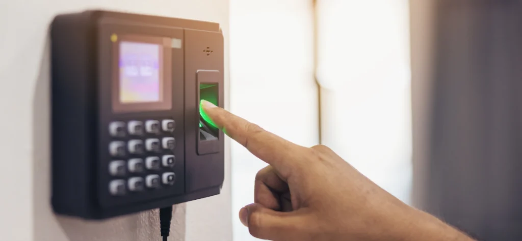 Uma mão pressionando o dedo indicador em um leitor biométrico de ponto eletrônico, que tem um teclado numérico e uma tela. O sensor biométrico está iluminado com uma luz verde.