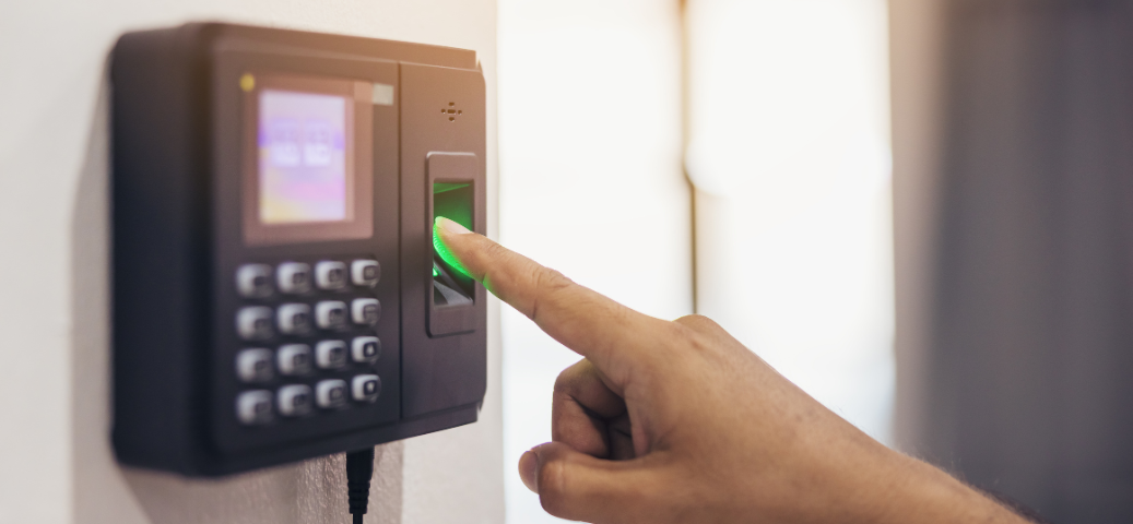 Uma mão pressionando o dedo indicador em um leitor biométrico de ponto eletrônico, que tem um teclado numérico e uma tela. O sensor biométrico está iluminado com uma luz verde.