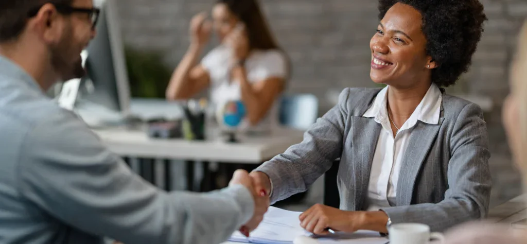Mulher negra sorridente de terno cinza apertando a mão de um colega em um escritório moderno e iluminado.