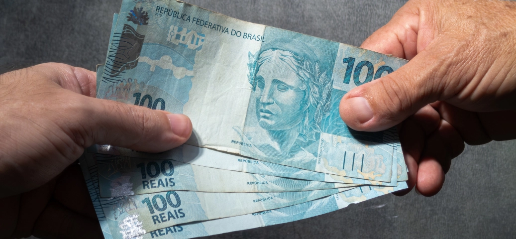 Duas mãos trocam notas de R$ 100 Reais, representando transação ou crédito fácil.