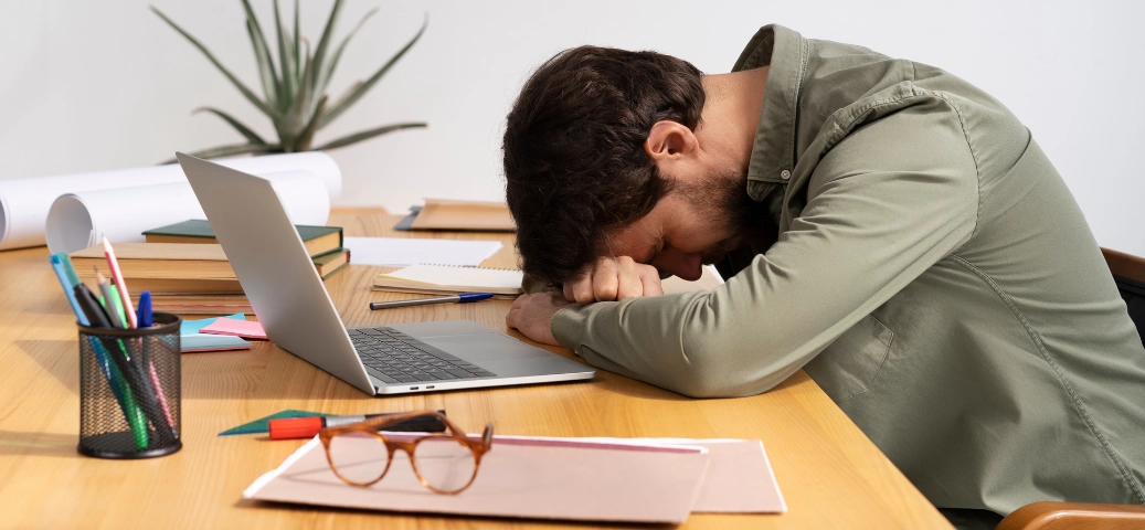 Homem com a cabeça apoiada sobre os braços em uma mesa de escritório com laptop, demonstrando cansaço ou exaustão.