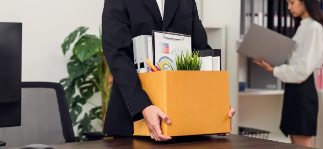 Pessoa vestindo terno e segurando uma caixa de papelão marrom, carregada com pertences de escritório como pastas, gráficos e um vaso pequeno. A cena se passa em um escritório e sugere uma situação de demissão ou mudança de emprego.