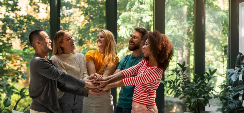 Cinco colegas de trabalho sorriem e unem as mãos em um gesto de colaboração em um escritório com vista para um jardim externo.