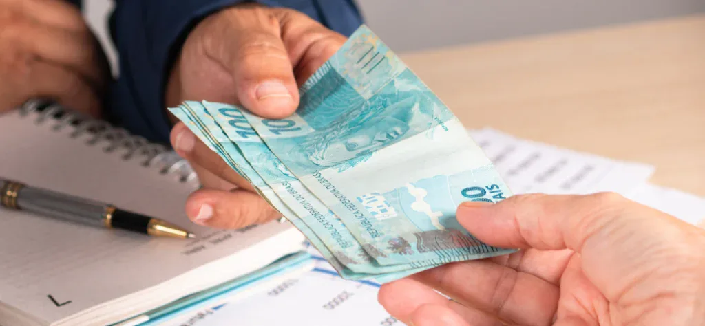 Close-up de uma mão entregando várias cédulas de cem reais para outra pessoa sobre uma mesa com caderno e caneta.