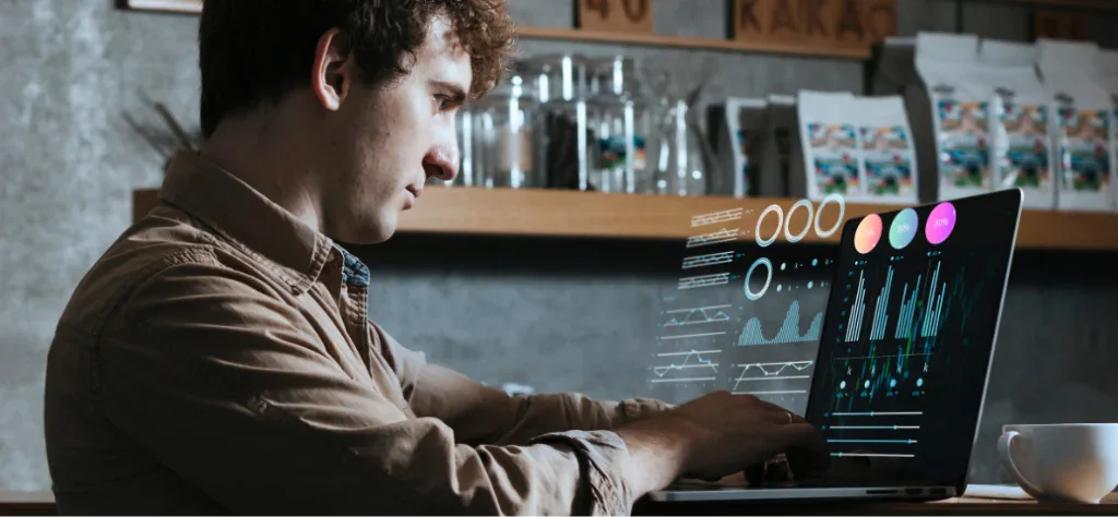Homem jovem em um café analisando gráficos e dados digitais holográficos que flutuam sobre a tela do seu notebook.