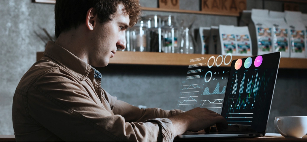 Homem jovem em um café analisando gráficos e dados digitais holográficos que flutuam sobre a tela do seu notebook.