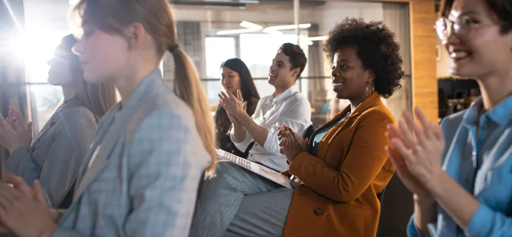 Público diversificado sentado em um auditório iluminado, sorrindo e aplaudindo durante um evento ou palestra corporativa.