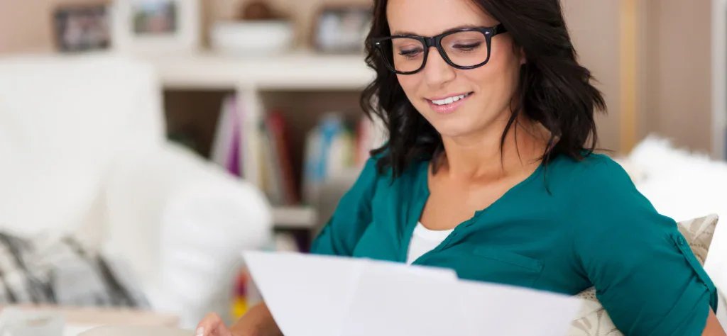 Uma mulher de óculos sorri ao analisar documentos financeiros, representando o planejamento para declarar o imposto de renda.