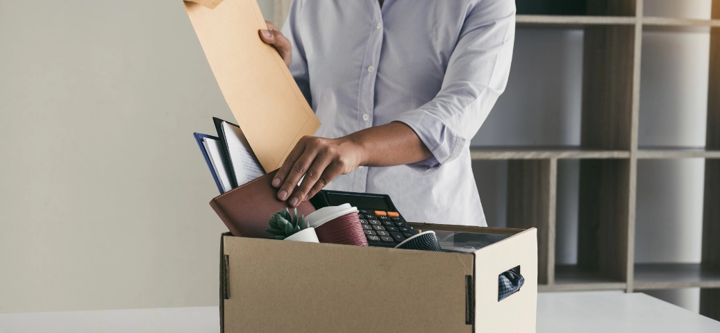 Pessoa de camisa lilás guarda pertences e uma agenda em uma caixa de papelão. Conceito de desligamento ou mudança.