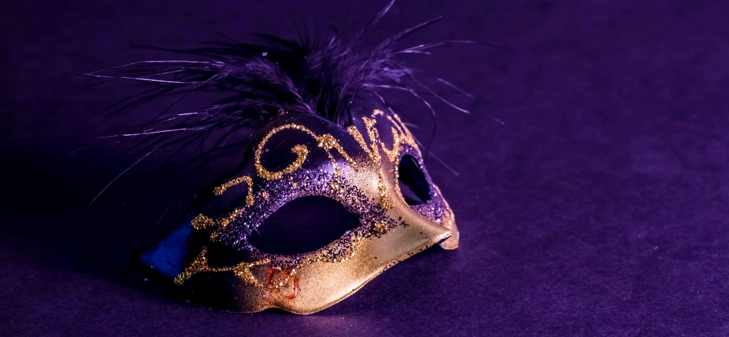 Máscara de Carnaval preta e dourada com glitter e penas, centralizada sobre um fundo roxo escuro e texturizado.