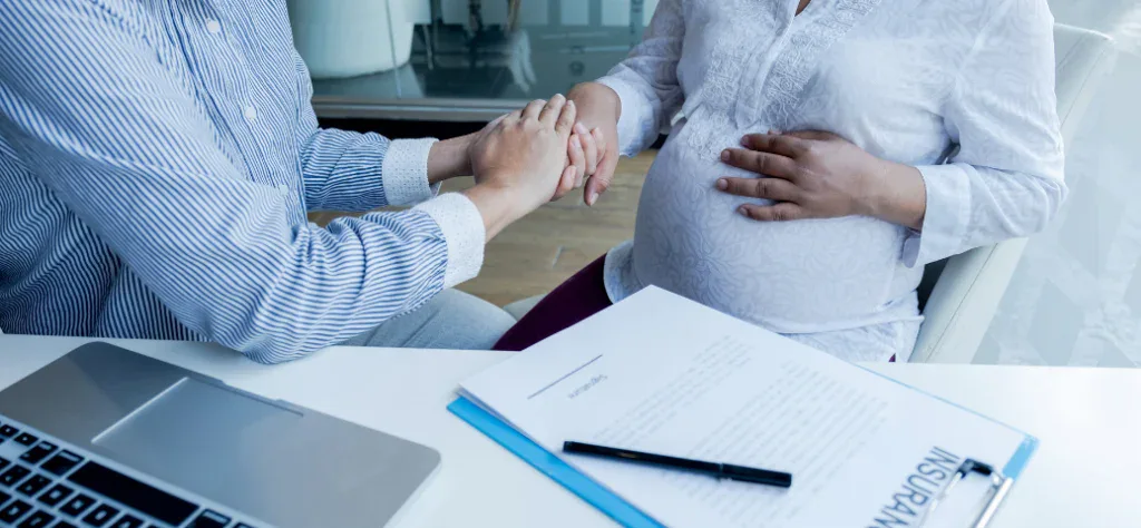 Uma agente de seguros segura as mãos de uma mulher grávida com a palavra "SEGURO" em um documento na mesa.