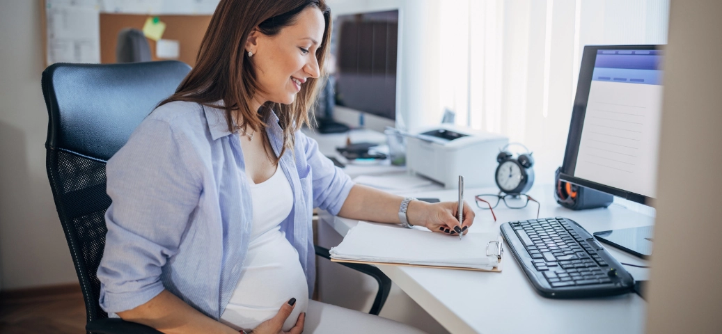 Mulher grávida sorridente escrevendo em uma prancheta em uma mesa de escritório, segurando a barriga.