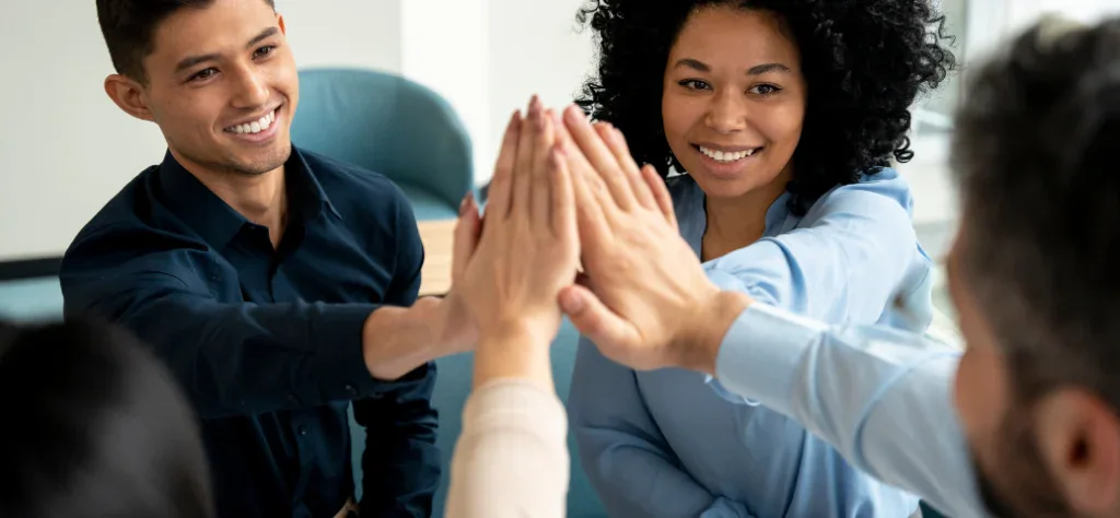 Equipe de trabalho diversa celebra o sucesso com um "high five" coletivo, demonstrando união, motivação e boa gestão de pessoas.