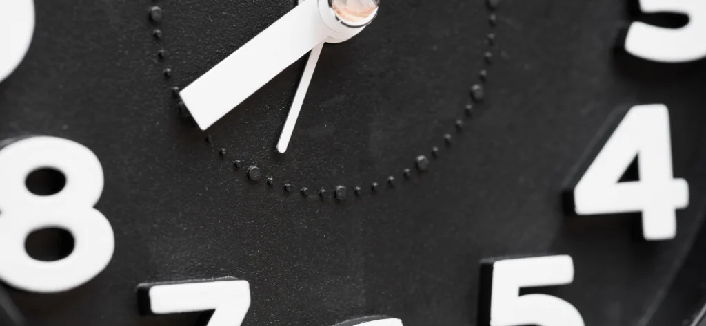 Close-up de um relógio analógico de parede preto com números brancos grandes, focando nos ponteiros das horas e minutos.