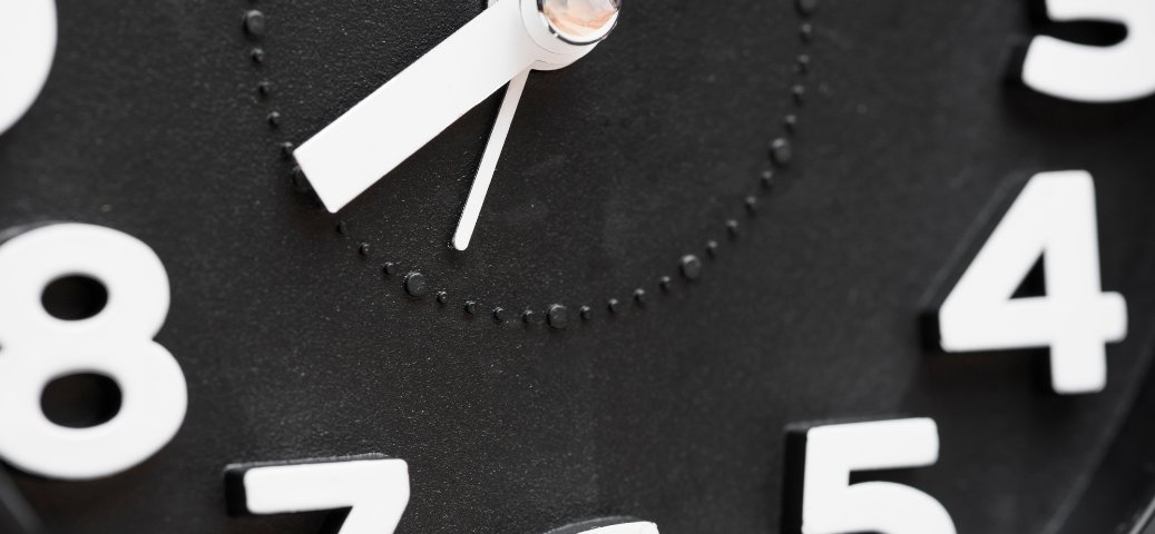 Close-up de um relógio analógico de parede preto com números brancos grandes, focando nos ponteiros das horas e minutos.
