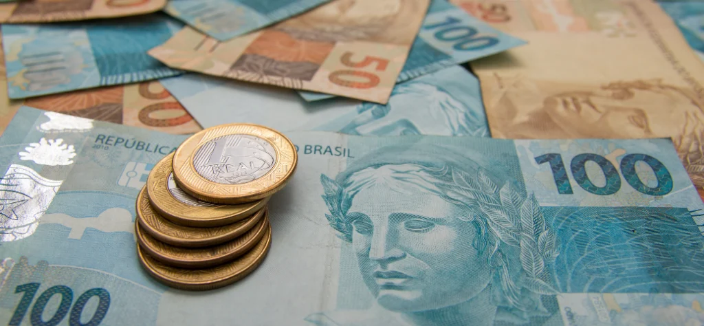 Moedas de 1 real sobre notas brasileiras de 100 e 50 reais espalhadas.