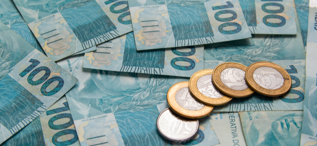 Várias cédulas de cem reais espalhadas sob quatro moedas de um real e uma de cinquenta centavos.