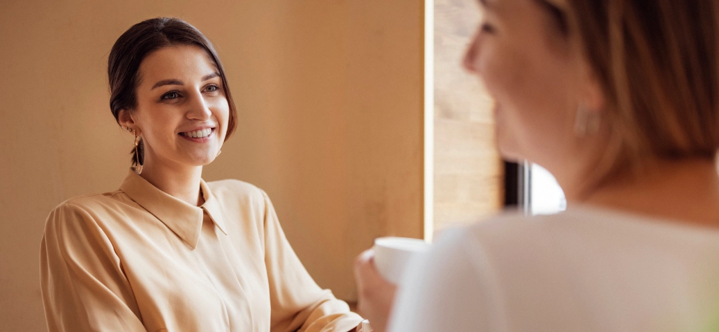 A imagem mostra duas mulheres em um ambiente interno iluminado e acolhedor, sugerindo uma conversa amigável ou profissional, como uma reunião de negócios casual ou um atendimento ao cliente.