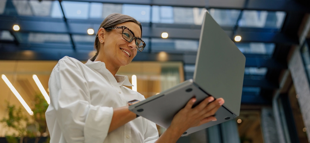 Mulher sorridente de óculos usando notebook em ambiente moderno com teto de vidro e iluminação decorativa ao fundo.