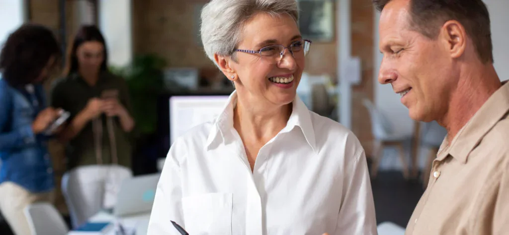 Mulher madura de óculos e camisa branca sorri enquanto conversa com um colega em um ambiente de escritório dinâmico.
