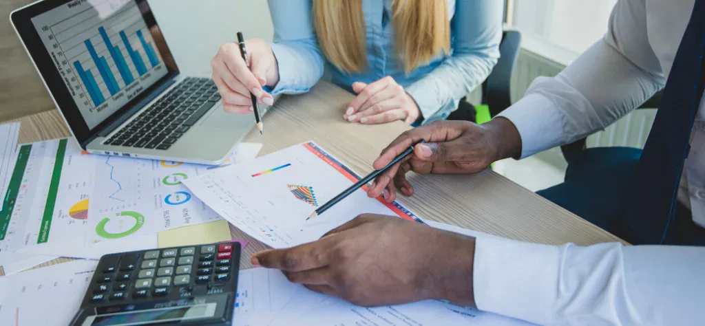 Mãos de duas pessoas analisando gráficos impressos e planilhas em um notebook, com uma calculadora sobre a mesa.