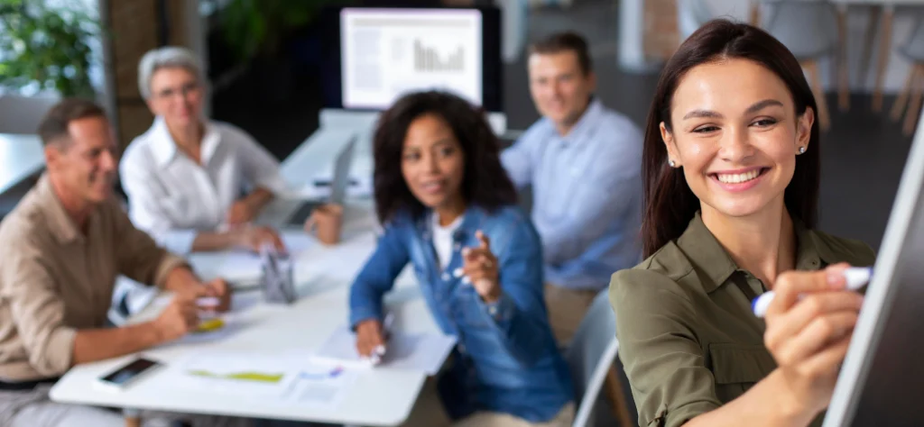 Mulher sorridente escrevendo em quadro branco, com equipe diversificada em reunião ao fundo no escritório.