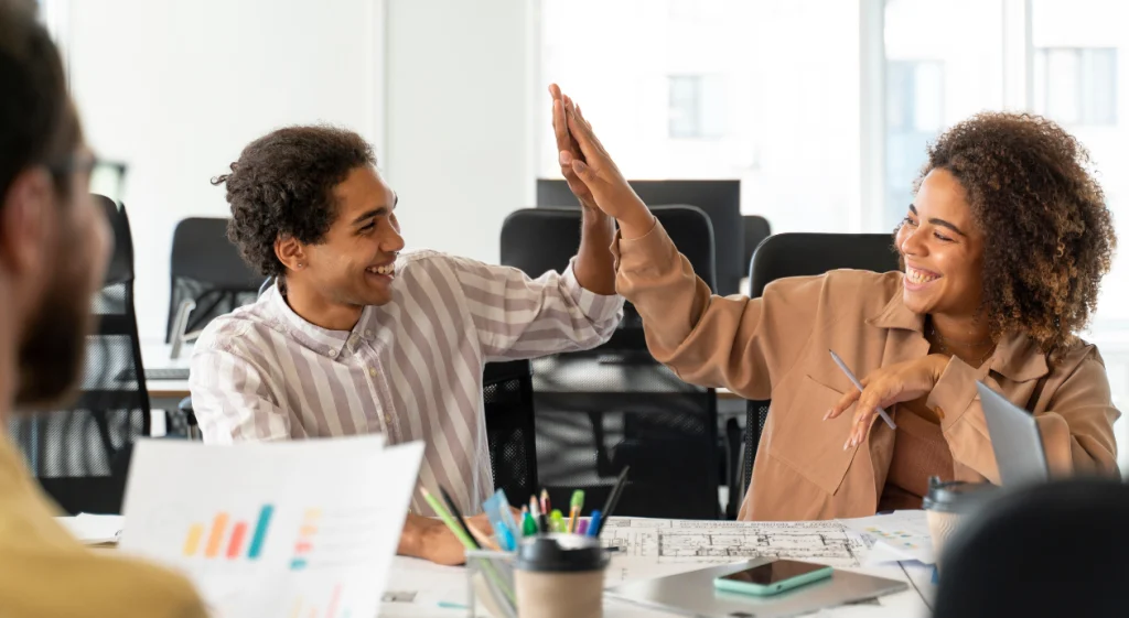 Dois colegas de trabalho sorridentes celebram com um "high five" (toca aqui) em uma mesa de escritório com projetos técnicos.