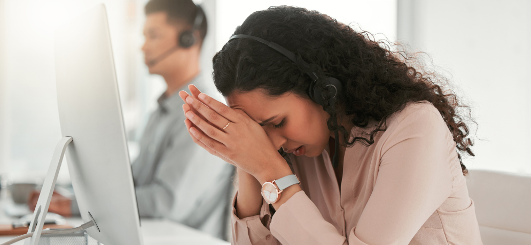 Mulher com fones de ouvido em um escritório, com as mãos no rosto e expressão de estresse ou cansaço profundo.
