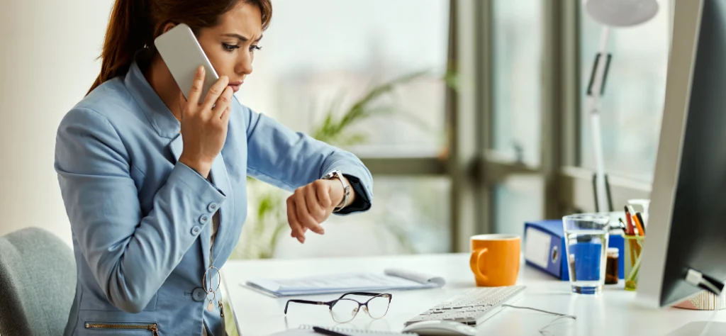 Mulher de blazer azul em um escritório, falando ao celular com expressão preocupada enquanto olha para o relógio de pulso.