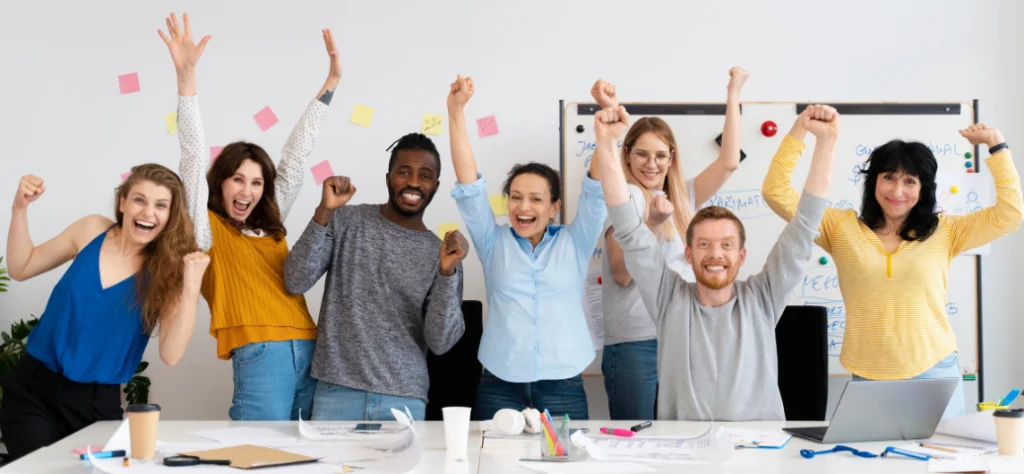 Grupo diverso de profissionais sorridentes comemora com braços erguidos em um escritório moderno e iluminado.