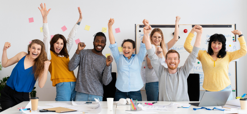 Grupo diverso de profissionais sorridentes comemora com braços erguidos em um escritório moderno e iluminado.