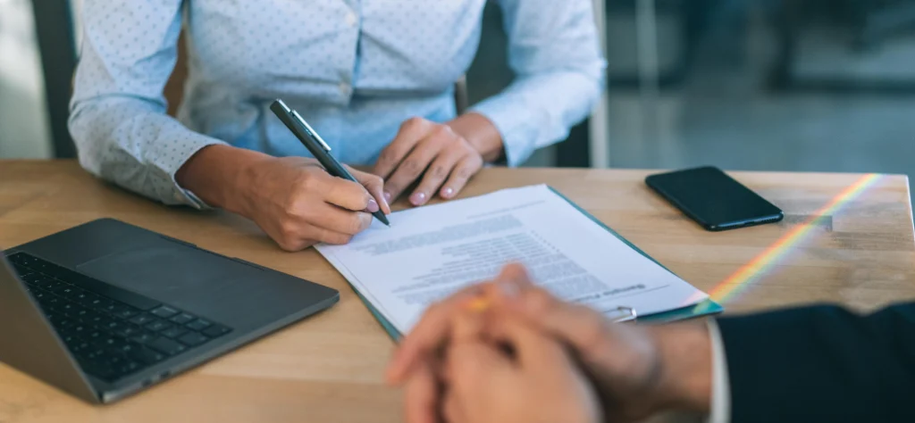 Pessoa com camisa azul clara assinando contrato sobre mesa de madeira, com notebook e celular ao lado em ambiente de escritório.