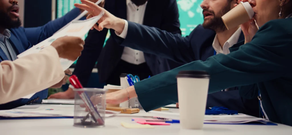 A imagem captura uma reunião de negócios intensa, com foco em uma mesa cheia de documentos, copos de café e materiais de escritório.