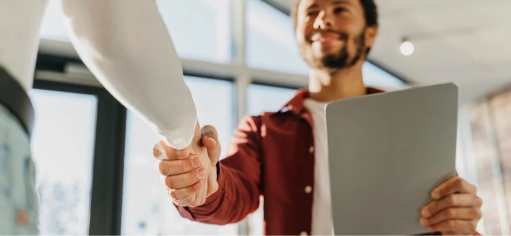Homem sorrindo segura tablet e aperta a mão de outra pessoa em escritório iluminado pelo sol durante reunião profissional.